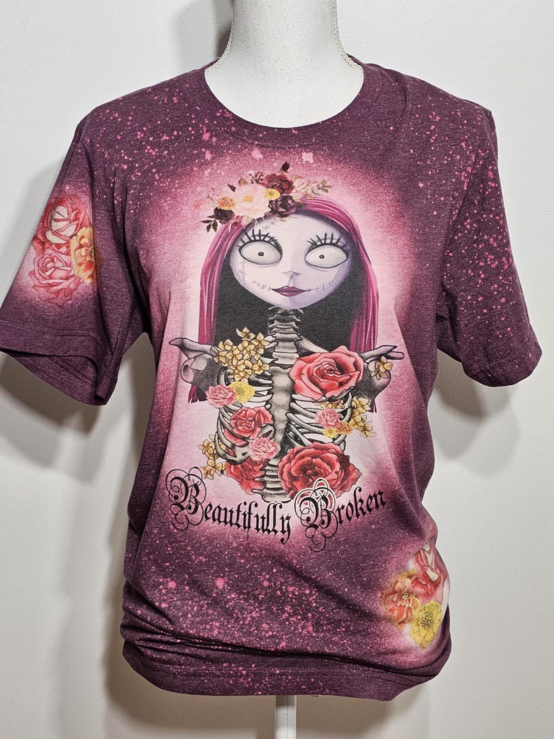 以下が含まれることがあります： 白とピンクの漂白剤でデザインされた、マロン色のTシャツ。骸骨に花が飾られ、「Beautifully Broken」という文字が書かれている。