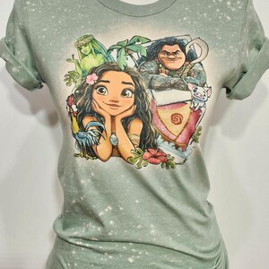 May include: Sage green t-shirt with a bleach-splattered effect. The shirt features a cartoon illustration of Moana, Maui, Pua, and a green-skinned woman with a flower crown. The text "Sage" is printed in white at the bottom of the image.