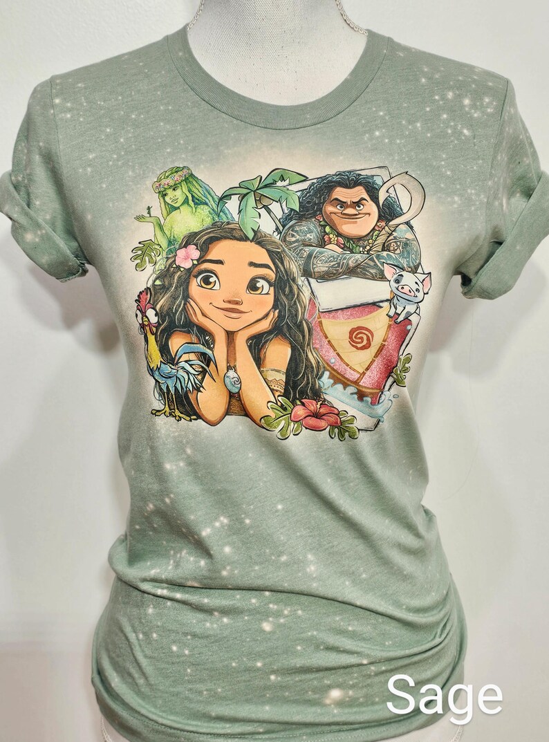 May include: Sage green t-shirt with a bleach-splattered effect. The shirt features a cartoon illustration of Moana, Maui, Pua, and a green-skinned woman with a flower crown. The text "Sage" is printed in white at the bottom of the image.