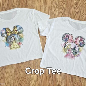 Peut inclure: Deux t-shirts courts blancs avec des motifs sur le th&egrave;me des princesses Disney. L'un repr&eacute;sente Belle, l'autre Aurore, dans une silhouette d'oreille de Mickey Mouse. Le texte "Crop Tee" est affich&eacute; en dessous.