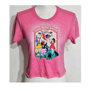 May include: Pink cropped t-shirt with a graphic of three girls in front of a tombstone with a flying Easy-Bake Oven and the text "Easy-Bake Coven".
