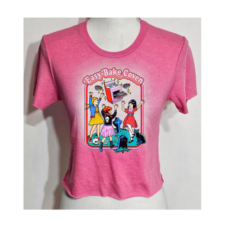 May include: Pink cropped t-shirt with a graphic of three girls in front of a tombstone with a flying Easy-Bake Oven and the text "Easy-Bake Coven".