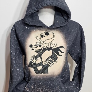 以下が含まれることがあります： 白い漂白剤が飛び散ったデザインの濃いグレーのフード付きスウェットシャツ。デザインは、ナイトメア・ビフォア・クリスマスのジャック・スケリントンとゼロの白黒のイラストです。