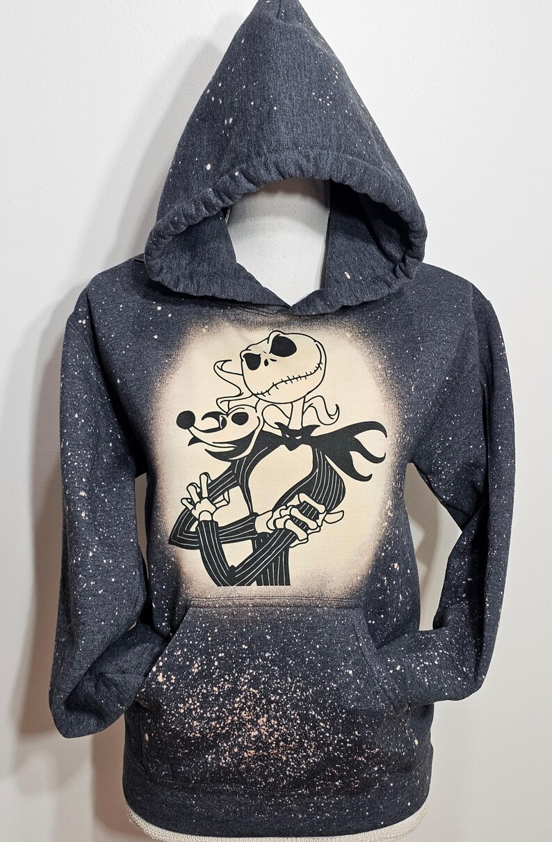 以下が含まれることがあります： 白い漂白剤が飛び散ったデザインの濃いグレーのフード付きスウェットシャツ。デザインは、ナイトメア・ビフォア・クリスマスのジャック・スケリントンとゼロの白黒のイラストです。