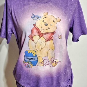 May include: A purple short-sleeve t-shirt with a bleached effect featuring a graphic of Winnie the Pooh, a blue bird, a butterfly, and a blue honey pot labeled Hunny. The text Pooh Bear is written in a cursive script below the character.