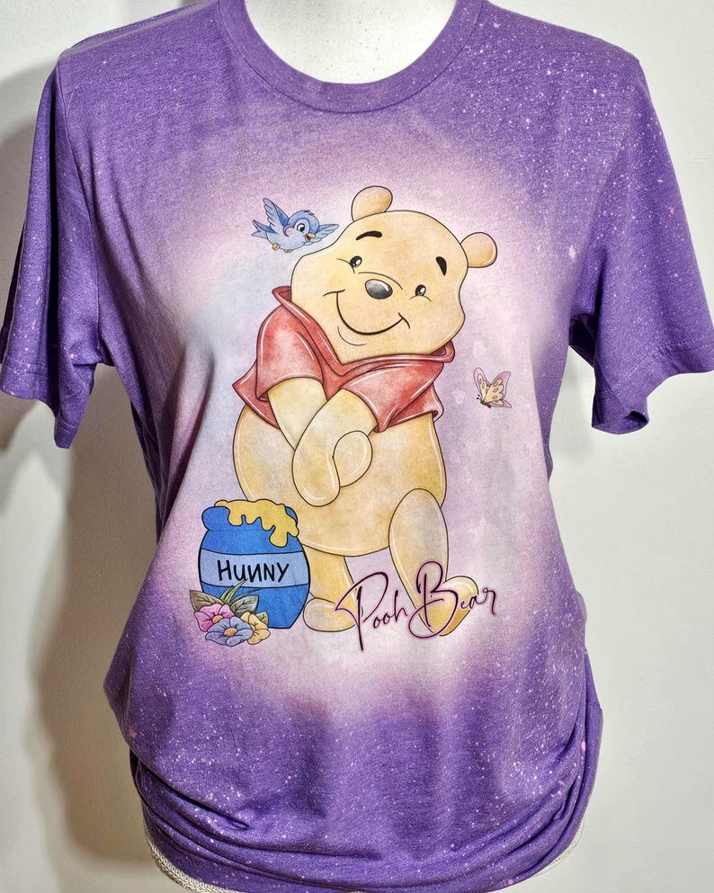 May include: A purple short-sleeve t-shirt with a bleached effect featuring a graphic of Winnie the Pooh, a blue bird, a butterfly, and a blue honey pot labeled Hunny. The text Pooh Bear is written in a cursive script below the character.