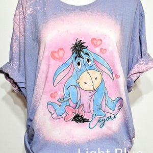 May include: A light blue bleached t-shirt featuring a graphic of Eeyore sitting with a pink bow and hearts. The design includes the name Eeyore in cursive script. The shirt has a distressed, mottled appearance from the bleaching process.