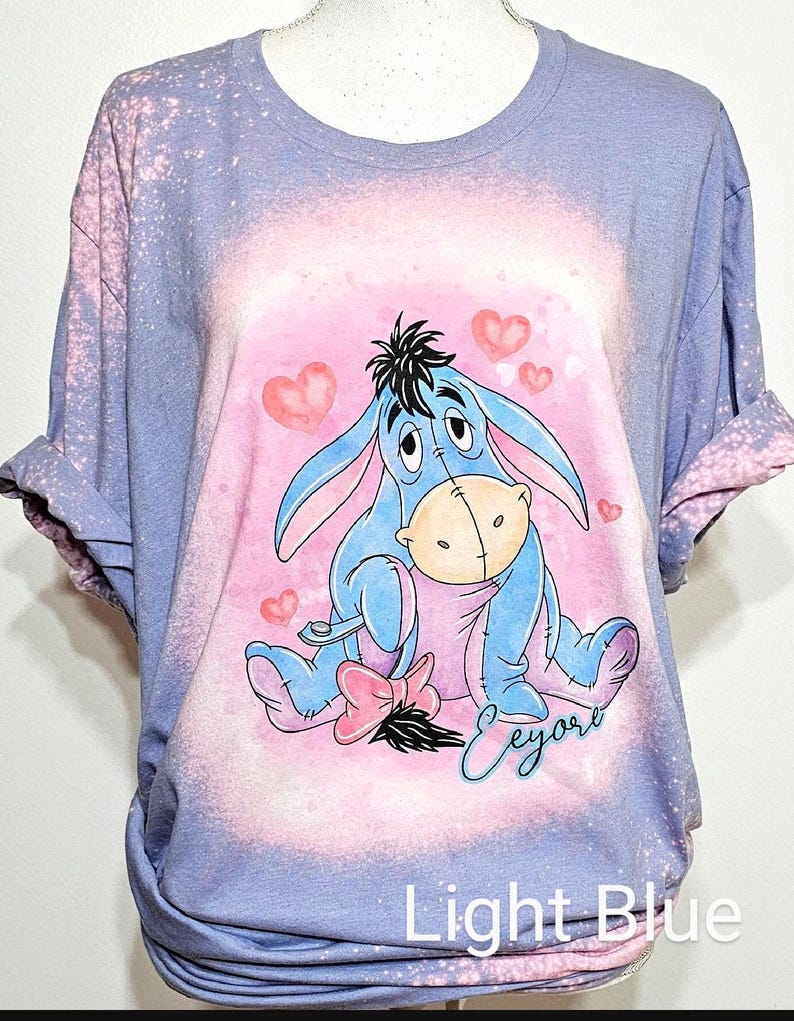 May include: A light blue bleached t-shirt featuring a graphic of Eeyore sitting with a pink bow and hearts. The design includes the name Eeyore in cursive script. The shirt has a distressed, mottled appearance from the bleaching process.