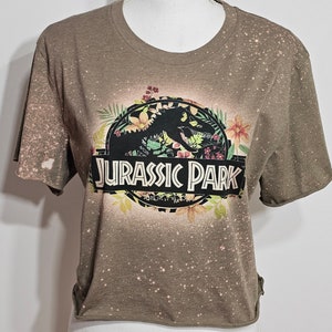 May include: A brown short-sleeved t-shirt with a bleached, speckled pattern throughout. The front features a circular graphic with a black dinosaur silhouette, colourful floral accents, and the text JURASSIC PARK in bold white capital letters.