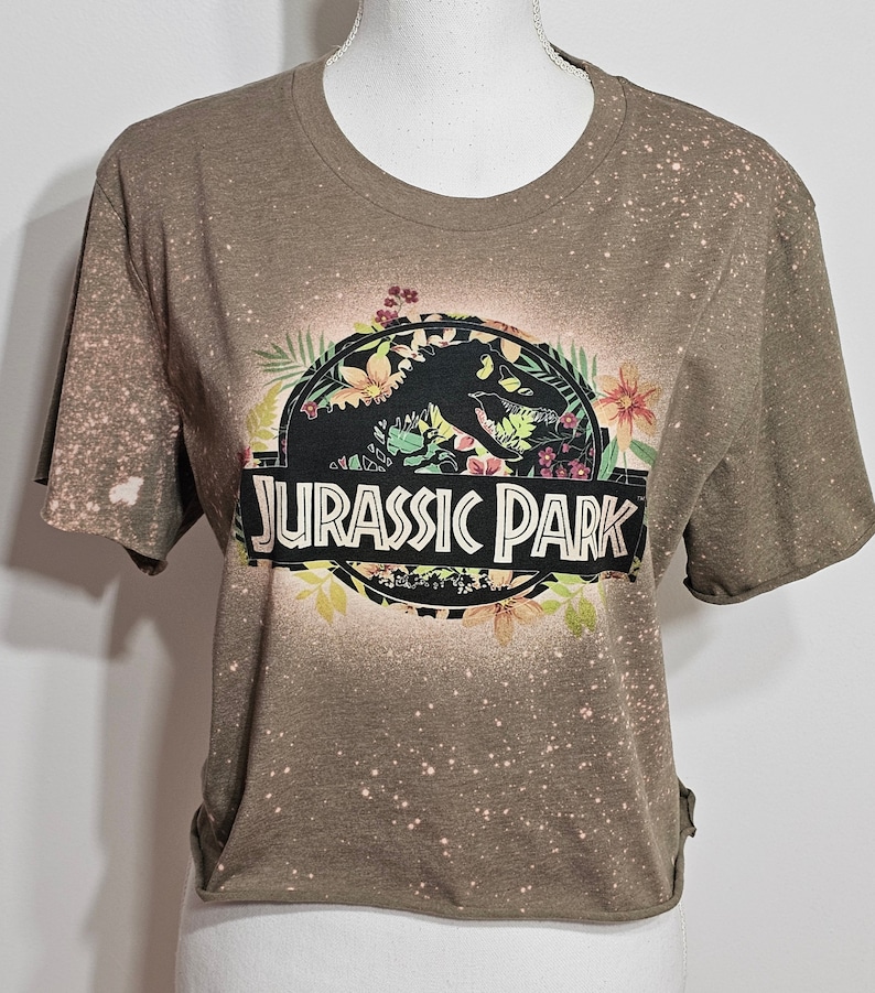 May include: A brown short-sleeved t-shirt with a bleached, speckled pattern throughout. The front features a circular graphic with a black dinosaur silhouette, colourful floral accents, and the text JURASSIC PARK in bold white capital letters.