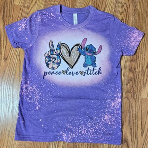May include: Purple tie-dye t-shirt with a peace sign, heart, and Stitch from Lilo & Stitch. The text reads "peace love stitch".