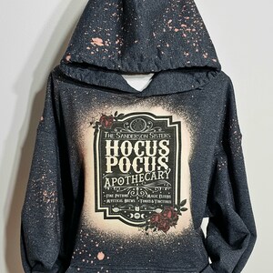 Puede incluir: Una sudadera con capucha negra con un dise&ntilde;o salpicado de lej&iacute;a. La parte delantera presenta una etiqueta de estilo vintage con el texto "The Sanderson Sisters Hocus Pocus Apothecary" y una lista de ingredientes: "Fine Potions, Magic Elixirs, Mystical Brews, Tonics & Tinctures".