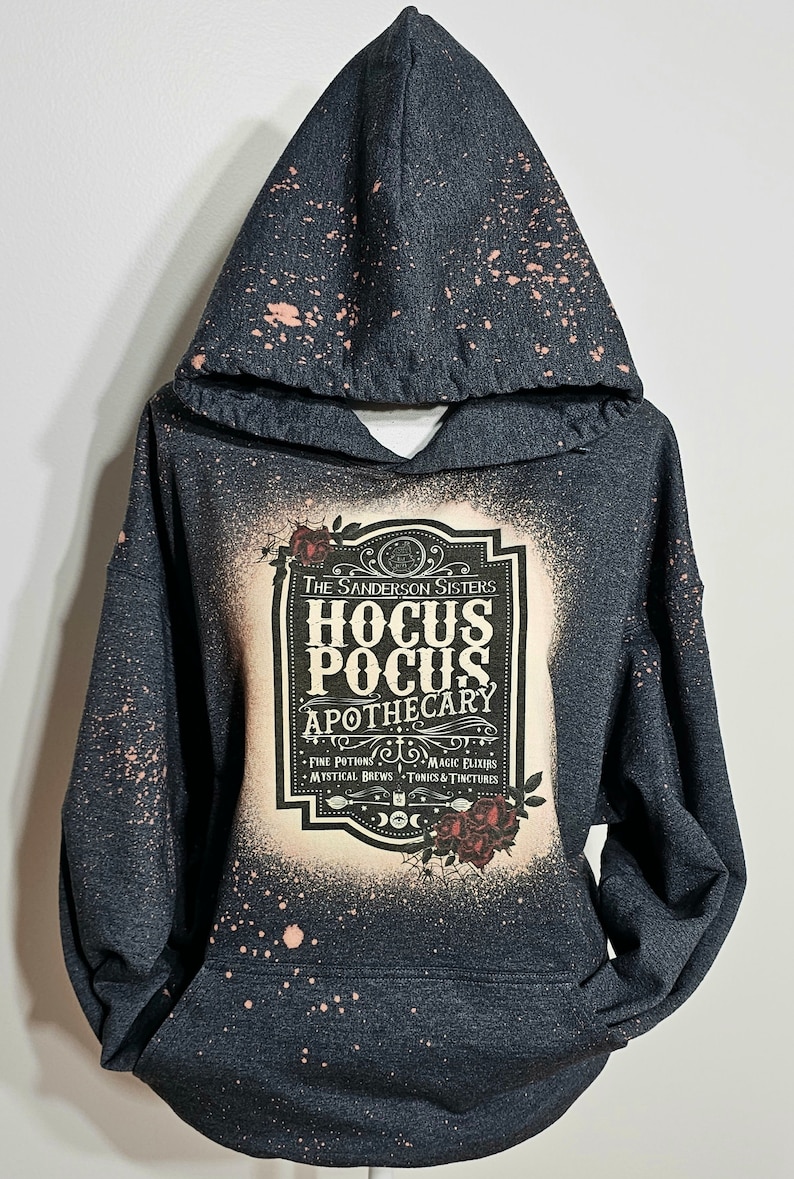 Puede incluir: Una sudadera con capucha negra con un dise&ntilde;o salpicado de lej&iacute;a. La parte delantera presenta una etiqueta de estilo vintage con el texto "The Sanderson Sisters Hocus Pocus Apothecary" y una lista de ingredientes: "Fine Potions, Magic Elixirs, Mystical Brews, Tonics & Tinctures".