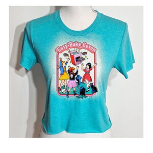 May include: A turquoise blue cropped t-shirt with a graphic print of four girls in front of an Easy-Bake Oven. The text "Easy-Bake Coven" is printed above the oven. The girls are wearing pink and black dresses and are holding up pies. The oven is pink and has a black top. The background is white with a faded effect.