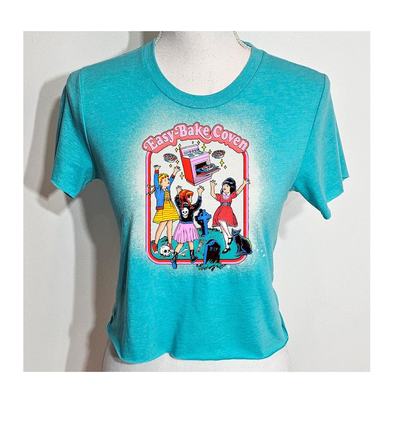 May include: A turquoise blue cropped t-shirt with a graphic print of four girls in front of an Easy-Bake Oven. The text "Easy-Bake Coven" is printed above the oven. The girls are wearing pink and black dresses and are holding up pies. The oven is pink and has a black top. The background is white with a faded effect.