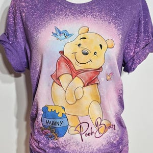 May include: A purple bleached t-shirt featuring a graphic of Winnie the Pooh in a red shirt, a blue honey pot labeled Hunny, a blue bird, and a butterfly. The script text Pooh Bear is printed in pink near the bottom right of the graphic design.