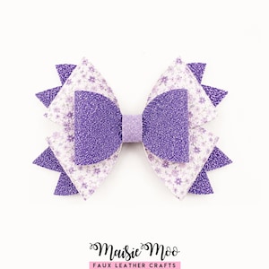 May include: A layered hair bow featuring a central purple glitter section, a light purple glitter center band, and outer layers of white faux leather with a small purple floral pattern and pointed purple glitter accents. Text reads Maisie Moo.