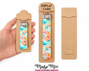 Pode incluir: Dois porta-chaves com um padrão abstrato colorido de formas azul-petróleo, laranja e rosa sobre um fundo branco, com ferragens de metal prateado e um anel, fixados em cartões de apresentação de cartão castanho com o texto DISPLAY CARD.