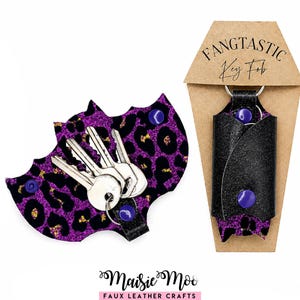 Puede incluir: Un llavero con forma de murciélago con estampado de leopardo morado y negro y un llavero de purpurina negra sobre una tarjeta de cartón con forma de ataúd etiquetada FANGTASTIC Key Fob, con botones de presión morados y anillas de metal.