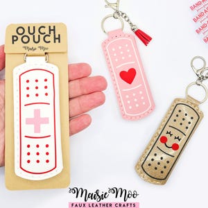 May include: Faux leather keychains in the shape of bandages. One is white with a pink cross, another is pink with a red heart, and the third is gold with a smiling face. The keychains have silver hardware and red tassels.