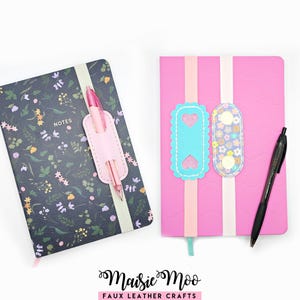 Könnte beinhalten: Ein dunkelblaues Notizbuch mit Blumenmuster und dem Wort NOTES, ausgestattet mit einem hellrosa Stiftehalter und einem rosa Stift. Daneben ein leuchtend rosa Notizbuch mit zwei Gummibändern, die ein blaues Herz-Etikett und ein Blumen-Etikett halten, sowie ein schwarzer Stift.