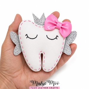 May include: A white faux leather tooth-shaped ornament featuring silver glitter wings and a silver glitter crown. The tooth has embroidered closed eyes, pink stitched edges, and a glossy pink bow. The text Maisie Moo Faux Leather Crafts is visible.