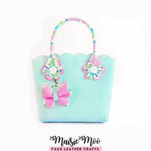 Può includere: Una borsa a mano azzurra con bordo smerlato, manico a motivo floreale e attacchi a forma di fiore. Un ciondolo a farfalla rosa pende da un lato. Il testo Maisie Moo FAUX LEATHER CRAFTS è stampato in nero e rosa sotto la borsa.