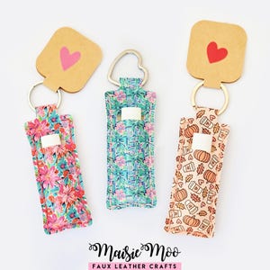 May include: Three lip balm holders with keyrings. One features a floral pattern with pink and red flowers, another has a blue and green floral design, and the third has a fall-themed pattern with pumpkins and coffee cups. Each has a cardboard tag with a heart.