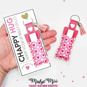 Op de afbeelding: Twee lippenbalsemhouders van roze kunstleer met een patroon van witte hartjes en stippen. Elke houder heeft een zilveren sleutelhanger, een lichtroze kwastje en een witte drukknoopsluiting. Eén houder zit vast aan een kaart met CHAPPY HUG.
