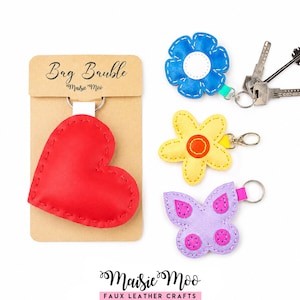Può includere: Una collezione di quattro portachiavi in similpelle che include un cuore rosso su un cartoncino espositore con l'etichetta Bag Bauble, un fiore blu, un fiore giallo con centro arancione e una farfalla viola chiaro con dettagli rosa, tutti dotati di anelli in metallo.