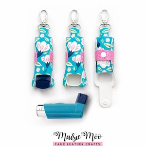Puede incluir: Tres fundas para inhalador de color verde azulado con un estampado floral en rosa, blanco y azul oscuro, cada una con una correa de purpurina rosa con cierre a presión y un broche giratorio plateado. Un inhalador se sitúa horizontalmente debajo.