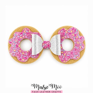Peut inclure: Un accessoire de coiffure composé de deux formes de beignets circulaires couleur beige avec un glaçage rose pailleté et des vermicelles colorés, reliés par un nœud central argenté pailleté. Le texte Maisie Moo FAUX LEATHER CRAFTS est imprimé en bas.