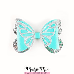 May include: A hair bow featuring a bright turquoise faux leather base layered with silver glitter accents. The design includes a cutout butterfly wing pattern in silver glitter over the turquoise centre, with additional silver glitter layers.