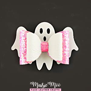 May include: A white ghost-shaped hair accessory featuring a layered bow made of white faux leather and bright pink glitter fabric. The ghost has two black oval eyes and a black oval mouth. The text Maisie Moo FAUX LEATHER CRAFTS is at the bottom.