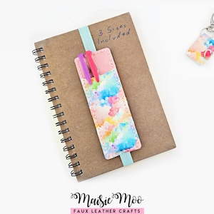 Op de afbeelding: Een bruin spiraalnotitieboek met een lichtblauwe elastische band die een rechthoekige kunstlederen pennenhouder met een veelkleurig aquarelpatroon vasthoudt. De houder bevat een paarse en een roze pen. Tekst leest 3 Sizes Included.