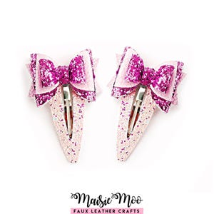 Puede incluir: Dos pinzas para el pelo con lazos superpuestos de purpurina rosa intenso y piel sintética rosa claro. Cada pinza tiene un cierre metálico plateado. El texto Maisie Moo FAUX LEATHER CRAFTS está impreso en negro en la parte inferior.