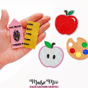 May include: A set of faux leather craft embellishments featuring a pink crayon, a yellow ruler, a red apple, a white glitter apple slice, and a wooden paint palette with colorful glitter paint spots. The text Maisie Moo Faux Leather Crafts is visible.