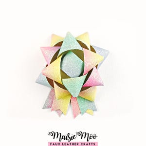 May include: A decorative bow crafted from glittery faux leather material featuring a pastel gradient of pink, yellow, and light blue. The bow has a multi-layered star shape with pointed loops and two hanging tails. Text below reads Maisie Moo FAUX LEATHER CRAFTS.