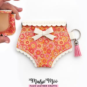 May include: A small pouch shaped like bloomers featuring a pink, orange, and red floral pattern with a white scalloped trim and a central white bow. It includes a silver keyring with a pink tassel. The text Maisie Moo Faux Leather Crafts is below.
