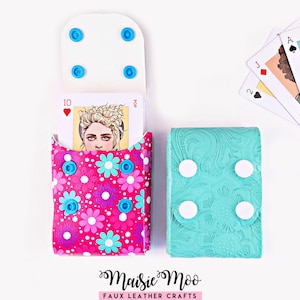 May include: Two rectangular card cases with snap closures. One case features a vibrant pink background with a multi-coloured floral pattern and holds a playing card with a portrait. The second case is solid turquoise with an embossed floral texture.