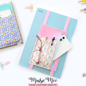 May include: A light blue 2025 planner with a pink elasticated organiser pouch attached to the front. The pouch features a white fabric with a colourful owl pattern, holding a white smartphone and a pair of glasses. A second organiser is visible nearby.