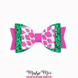 May include: A handmade bow featuring a strawberry pattern on white, green glitter accents, and a bright pink base. The bow is made of faux leather and has a textured finish. The text "Maisie Moo Faux Leather Crafts" is at the bottom.
