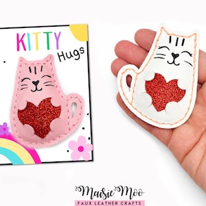 May include: A pink faux leather cat-shaped patch with a red glitter heart and a white faux leather cat-shaped patch with a red glitter heart, both featuring stitched edges and black ink facial details, displayed on a card labeled KITTY HUGS.