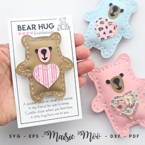 Könnte beinhalten: Drei bärenförmige Plüschornamente mit genähten Kanten. Ein beigefarbener Bär befindet sich auf einer weißen Karte mit der Aufschrift BEAR HUG und Text. Die anderen beiden sind hellblau mit einem Leopardenherz und hellrosa mit einem Blumenherz.