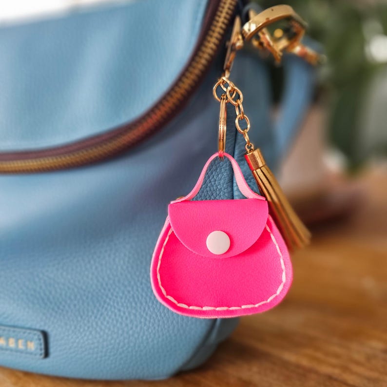 Peut inclure: Un porte-cl&eacute;s en forme de sac &agrave; main rose vif, avec un bouton blanc et des coutures blanches. Le porte-cl&eacute;s est suspendu &agrave; une cha&icirc;ne et un pompon dor&eacute;s, sur fond de sac &agrave; main bleu clair.