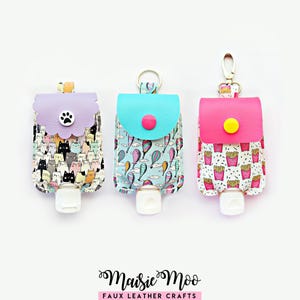 May include: Three rectangular faux leather hand sanitizer holders with metal keychains. The left features a cat print and lavender flap, the center has a hot air balloon print and turquoise flap, and the right has a french fry print and pink flap.