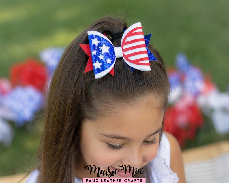 Op de afbeelding: Een patriottische haarstrik met rode, witte en blauwe kleuren. De strik heeft een wit midden, rood-witte strepen aan &eacute;&eacute;n kant en een blauw gedeelte met witte sterren aan de andere kant. Bevestigd met een zwarte haarband.