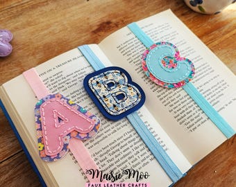 Op de afbeelding: Drie elastische boekenleggers met de letters A, B en C. De A is roze met een bloemenpatroon, de B is marineblauw met een bijenpatroon en de C is lichtblauw met een bloemenpatroon. Elke letter is vastgestikt op een elastische band.