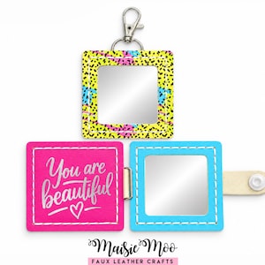 May include: A square folding keychain mirror with a silver metal clasp. One side features a yellow, pink, and blue speckled pattern. The interior displays a bright pink cover with the white text You are beautiful and a heart icon, plus a mirror.