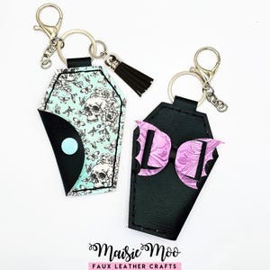 May include: Two coffin-shaped keychains. One features a teal background with skulls and floral patterns, the other is black with a pink bow and bat wings. Both have silver key rings and clasps, and one has a black tassel.